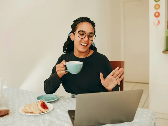 A woman at her kitchen table on a video call, laughing, a cup of coffee in her hand.